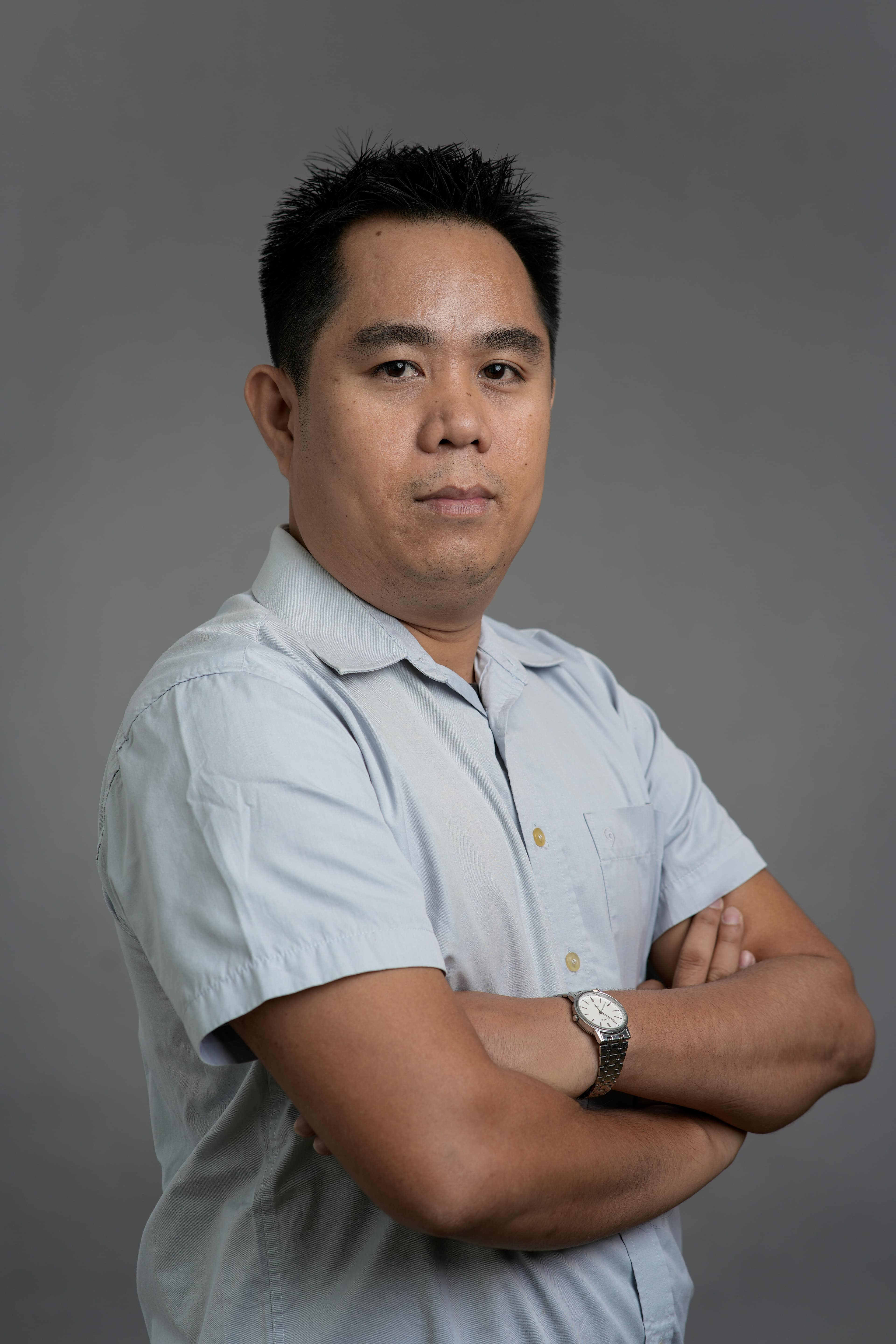 Romualdo Dasig standing with arms folded, in a pale blue short-sleeved shirt against a grey studio backdrop.